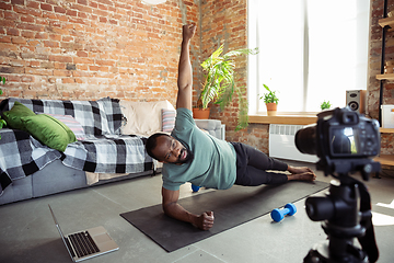 Image showing Young african-american man teaching at home online courses of fitness, aerobic, sporty lifestyle during quarantine, reording on camera, streaming