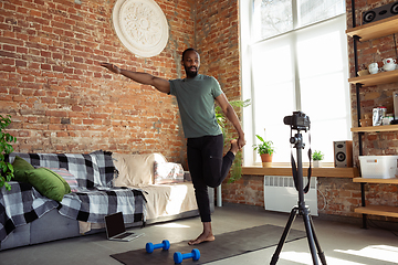 Image showing Young african-american man teaching at home online courses of fitness, aerobic, sporty lifestyle during quarantine, reording on camera, streaming