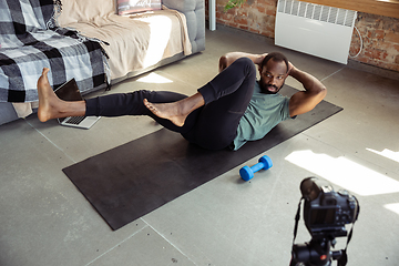 Image showing Young african-american man teaching at home online courses of fitness, aerobic, sporty lifestyle during quarantine, reording on camera, streaming