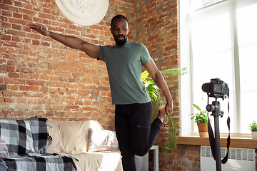 Image showing Young african-american man teaching at home online courses of fitness, aerobic, sporty lifestyle during quarantine, reording on camera, streaming