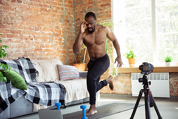 Image showing Young african-american man teaching at home online courses of fitness, aerobic, sporty lifestyle during quarantine, reording on camera, streaming