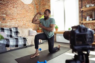 Image showing Young african-american man teaching at home online courses of fitness, aerobic, sporty lifestyle during quarantine, reording on camera, streaming