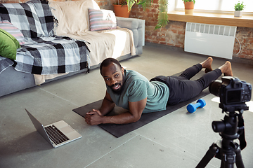 Image showing Young african-american man teaching at home online courses of fitness, aerobic, sporty lifestyle during quarantine, reording on camera, streaming