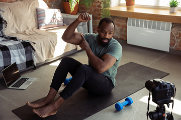 Image showing Young african-american man teaching at home online courses of fitness, aerobic, sporty lifestyle during quarantine, reording on camera, streaming