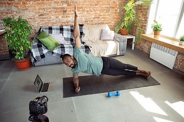 Image showing Young african-american man teaching at home online courses of fitness, aerobic, sporty lifestyle during quarantine, reording on camera, streaming