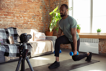 Image showing Young african-american man teaching at home online courses of fitness, aerobic, sporty lifestyle during quarantine, reording on camera, streaming