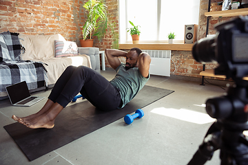 Image showing Young african-american man teaching at home online courses of fitness, aerobic, sporty lifestyle during quarantine, reording on camera, streaming