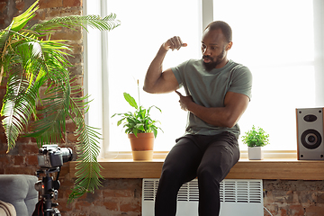 Image showing Young african-american man teaching at home online courses of fitness, aerobic, sporty lifestyle during quarantine, reording on camera, streaming