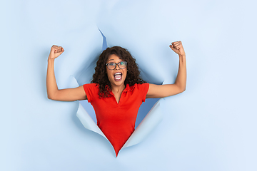 Image showing Cheerful young woman poses in torn blue paper hole background, emotional and expressive