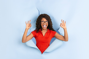 Image showing Cheerful young woman poses in torn blue paper hole background, emotional and expressive