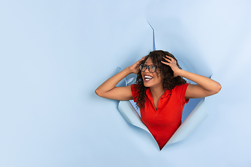 Image showing Cheerful young woman poses in torn blue paper hole background, emotional and expressive