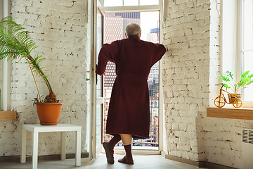 Image showing Mature senior older man during quarantine, realizing how important stay at home during virus outbreak, looking throught window on day Boston city