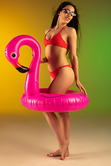 Image showing Fashion portrait of young fit and sportive woman in stylish red luxury swimwear with rubber flamingo on gradient background. Perfect body ready for summertime.