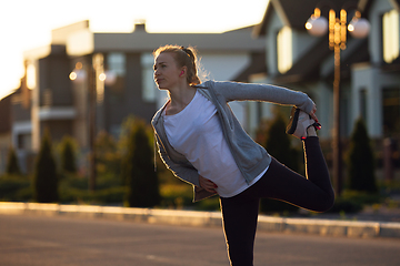 Image showing Young female runner, athlete is stretching before jogging in the city street in sunshine. Beautiful caucasian woman training, listening to music