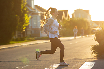 Image showing Young female runner, athlete is jogging in the city street in sunshine. Beautiful caucasian woman training, listening to music