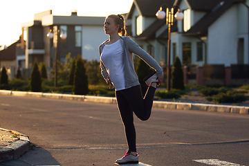 Image showing Young female runner, athlete is stretching before jogging in the city street in sunshine. Beautiful caucasian woman training, listening to music