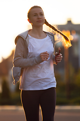 Image showing Young female runner, athlete is jogging in the city street in sunshine. Beautiful caucasian woman training, listening to music