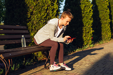 Image showing Young female runner, athlete resting after jogging in the city street in sunshine. Beautiful caucasian woman training, listening to music