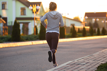 Image showing Young female runner, athlete is jogging in the city street in sunshine. Beautiful caucasian woman training, listening to music