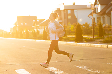 Image showing Young female runner, athlete is jogging in the city street in sunshine. Beautiful caucasian woman training, listening to music