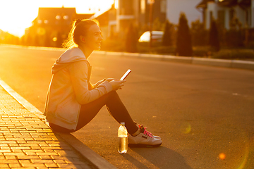 Image showing Young female runner, athlete resting after jogging in the city street in sunshine. Beautiful caucasian woman training, listening to music