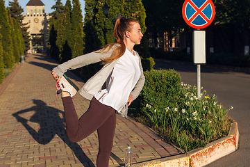 Image showing Young female runner, athlete is stretching before jogging in the city street in sunshine. Beautiful caucasian woman training, listening to music