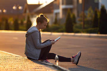 Image showing Young female runner, athlete resting after jogging in the city street in sunshine. Beautiful caucasian woman training, listening to music