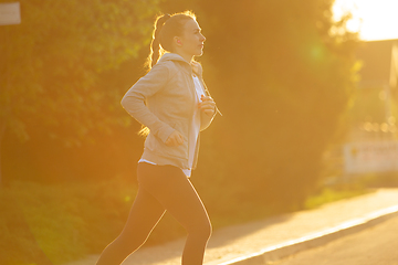 Image showing Young female runner, athlete is jogging in the city street in sunshine. Beautiful caucasian woman training, listening to music