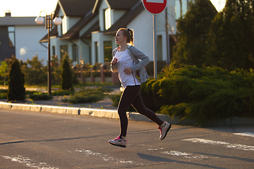 Image showing Young female runner, athlete is jogging in the city street in sunshine. Beautiful caucasian woman training, listening to music