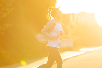 Image showing Young female runner, athlete is jogging in the city street in sunshine. Beautiful caucasian woman training, listening to music