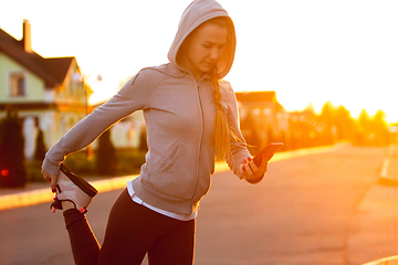 Image showing Young female runner, athlete is stretching before jogging in the city street in sunshine. Beautiful caucasian woman training, listening to music