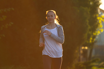 Image showing Young female runner, athlete is jogging in the city street in sunshine. Beautiful caucasian woman training, listening to music