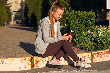 Image showing Young female runner, athlete resting after jogging in the city street in sunshine. Beautiful caucasian woman training, listening to music