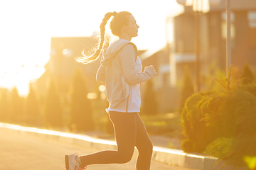 Image showing Young female runner, athlete is jogging in the city street in sunshine. Beautiful caucasian woman training, listening to music