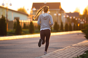 Image showing Young female runner, athlete is jogging in the city street in sunshine. Beautiful caucasian woman training, listening to music