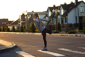 Image showing Young female runner, athlete is stretching before jogging in the city street in sunshine. Beautiful caucasian woman training, listening to music