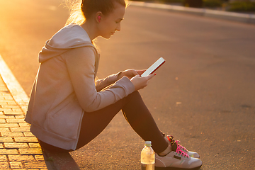 Image showing Young female runner, athlete resting after jogging in the city street in sunshine. Beautiful caucasian woman training, listening to music