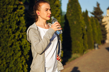 Image showing Young female runner, athlete resting after jogging in the city street in sunshine. Beautiful caucasian woman training, listening to music