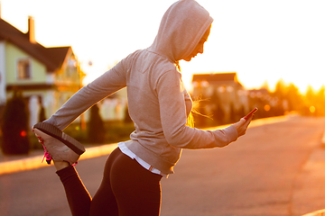 Image showing Young female runner, athlete is stretching before jogging in the city street in sunshine. Beautiful caucasian woman training, listening to music