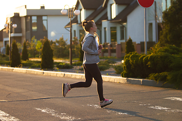 Image showing Young female runner, athlete is jogging in the city street in sunshine. Beautiful caucasian woman training, listening to music