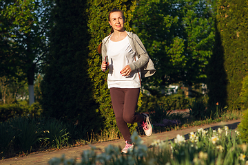 Image showing Young female runner, athlete is jogging in the city street in sunshine. Beautiful caucasian woman training, listening to music