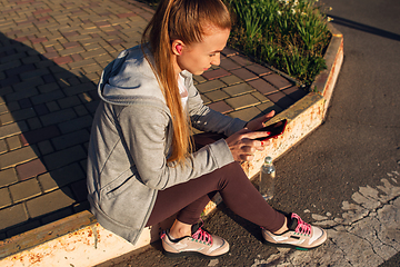 Image showing Young female runner, athlete resting after jogging in the city street in sunshine. Beautiful caucasian woman training, listening to music