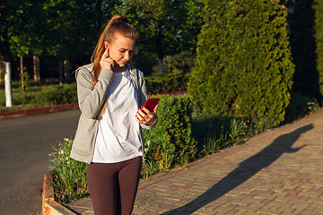 Image showing Young female runner, athlete resting after jogging in the city street in sunshine. Beautiful caucasian woman training, listening to music