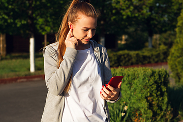 Image showing Young female runner, athlete resting after jogging in the city street in sunshine. Beautiful caucasian woman training, listening to music