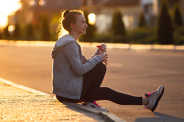 Image showing Young female runner, athlete resting after jogging in the city street in sunshine. Beautiful caucasian woman training, listening to music