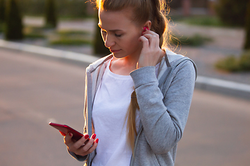 Image showing Young female runner, athlete resting after jogging in the city street in sunshine. Beautiful caucasian woman training, listening to music