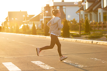 Image showing Young female runner, athlete is jogging in the city street in sunshine. Beautiful caucasian woman training, listening to music