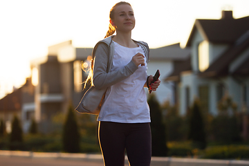 Image showing Young female runner, athlete is jogging in the city street in sunshine. Beautiful caucasian woman training, listening to music