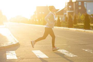 Image showing Young female runner, athlete is jogging in the city street in sunshine. Beautiful caucasian woman training, listening to music