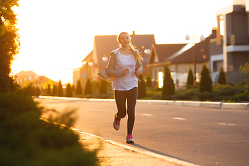 Image showing Young female runner, athlete is jogging in the city street in sunshine. Beautiful caucasian woman training, listening to music